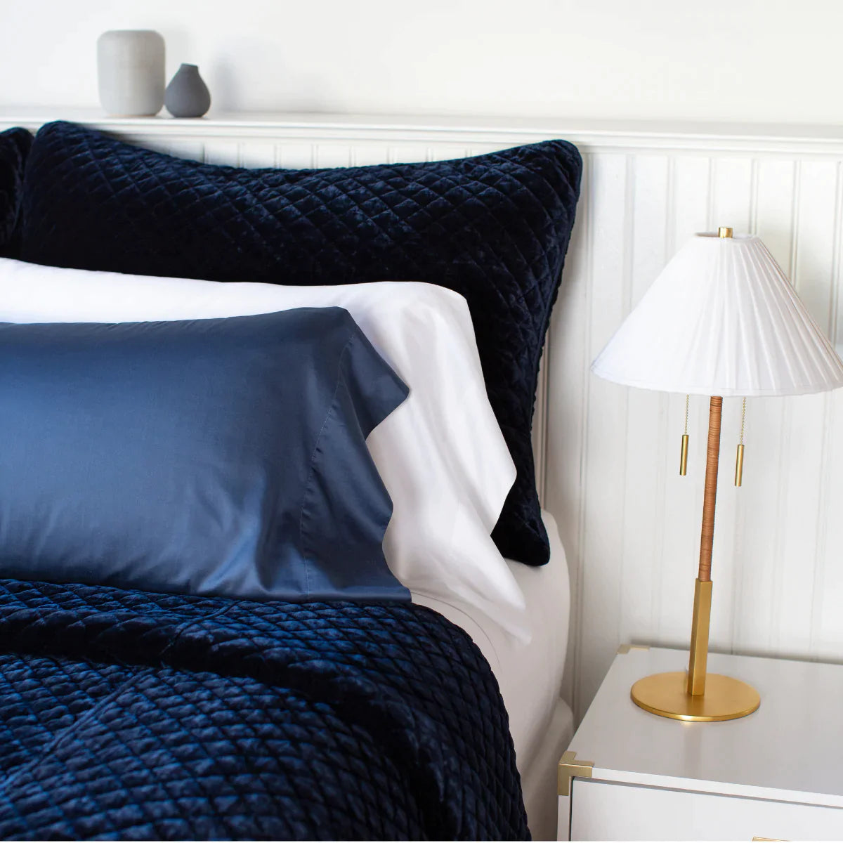 : White Bria fitted sheet on a neatly made bed shown from close up three quarter angle. Layered with deep blue silk velvet quilted shams and coverlet, it showcases a minimalist and gender neutral look.