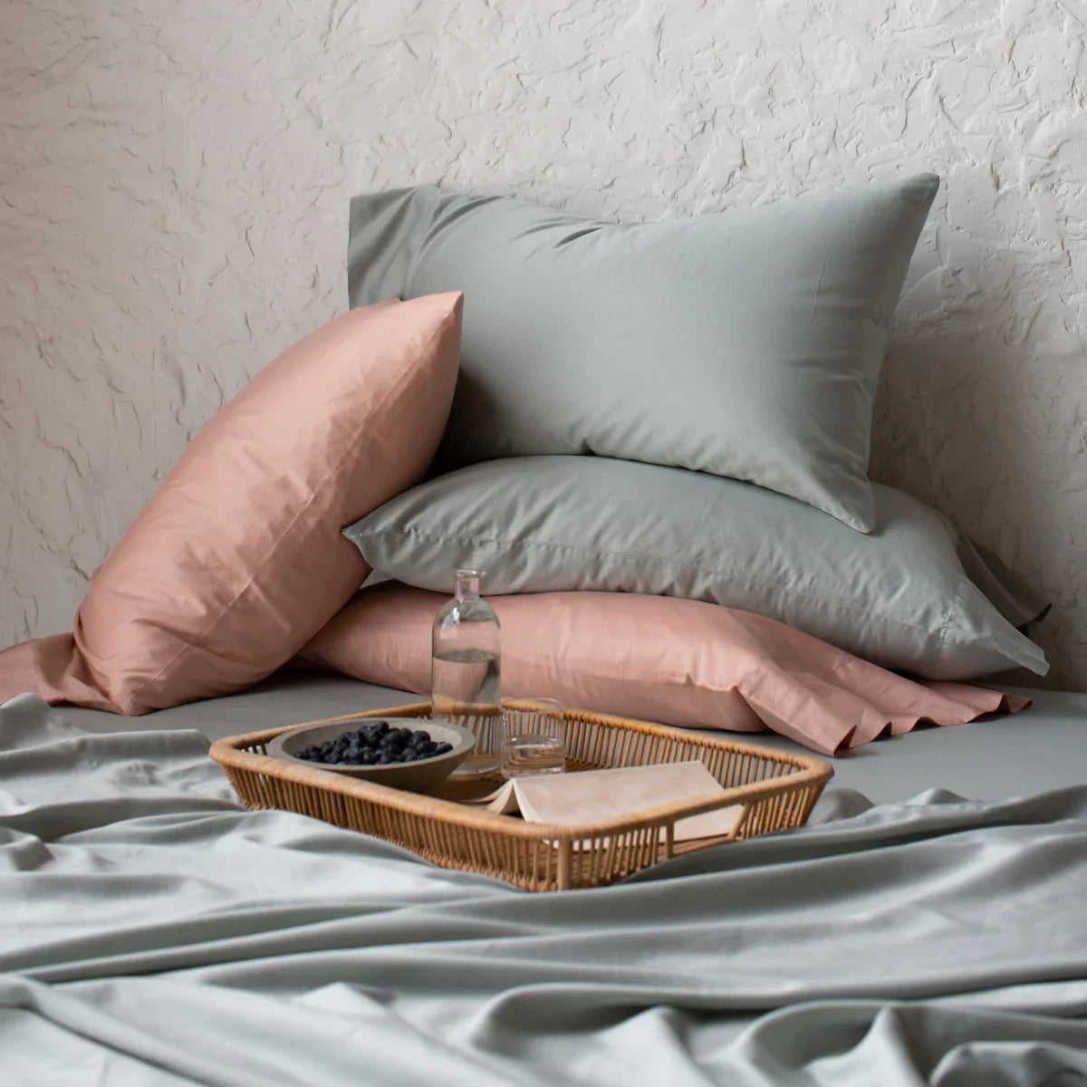 : Four Bria sleeping pillows in soft green and pink tones, stacked haphazardly on a matching bed against a textured white wall, perfect for lounging while enjoying the fruit and open book on the wicker tray.