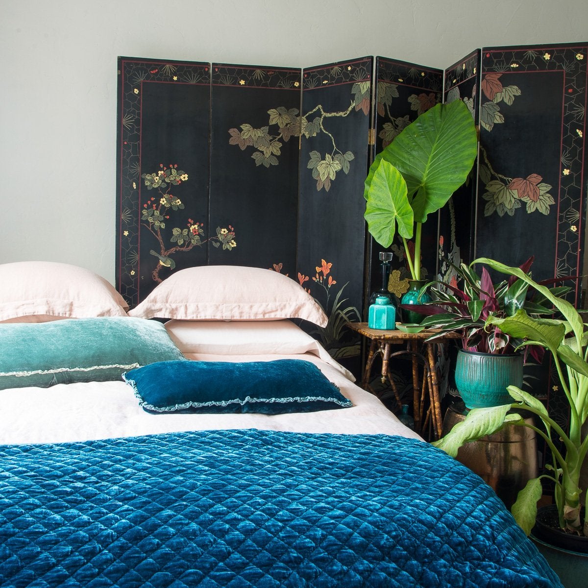 : On a pearl pink linen bed, Carmen throw pillows in contrasting blue-green tones lay flat, highlighting the charmeuse, petite, raw-edge ruffle. Styled with on-tone ceramics and big green plants in front of a dark floral room divider.