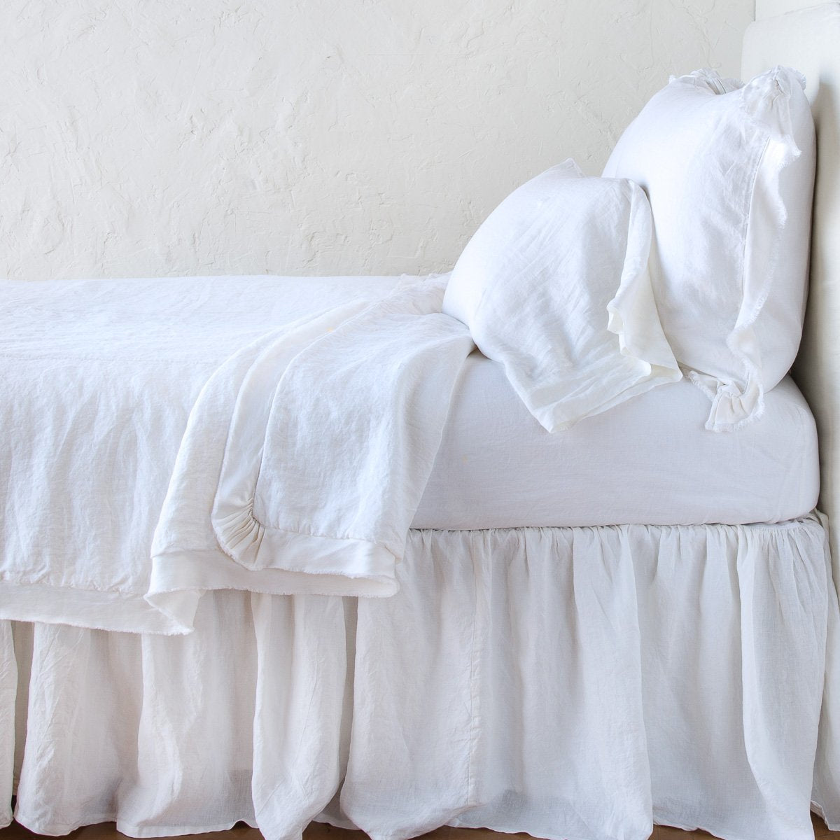 White: satin trimmed linen coverlet on monochromatic bed with corner pulled back to reveal sheet, bed skirt - side view.