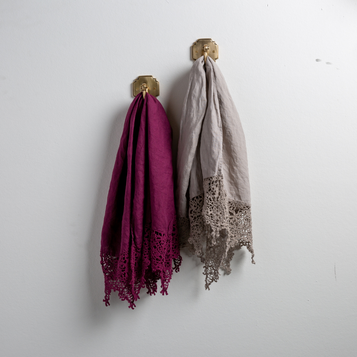 : Two Frida guest towels, draped over decorative hooks against a white wall - fig and fog.