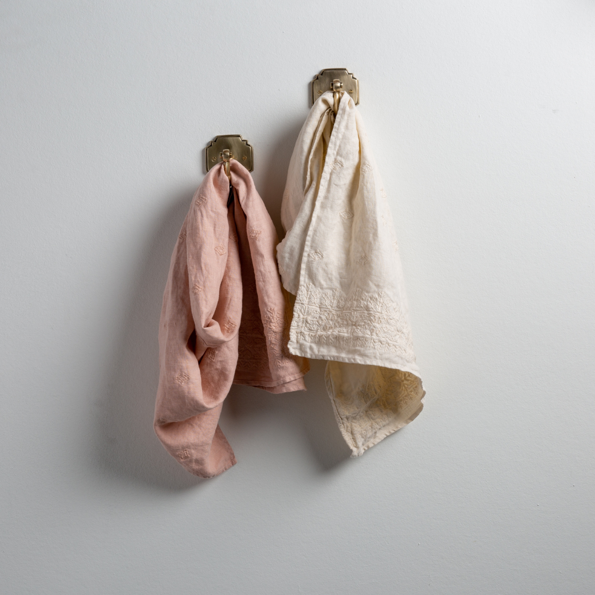 : Two Ines guest towels, draped over decorative hooks against a white wall - rouge and parchment.
