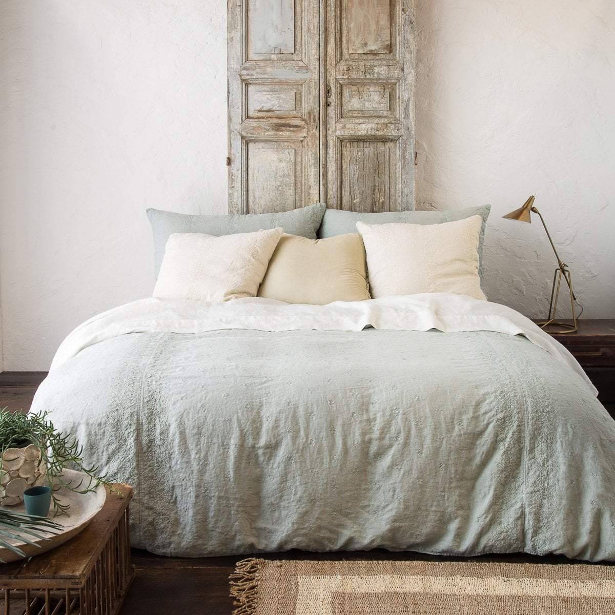 : Ines duvet cover in eucalyptus with cream-toned sheets and pillows, in a room with distressed wood, light walls, and neutral accents - end of bed view.