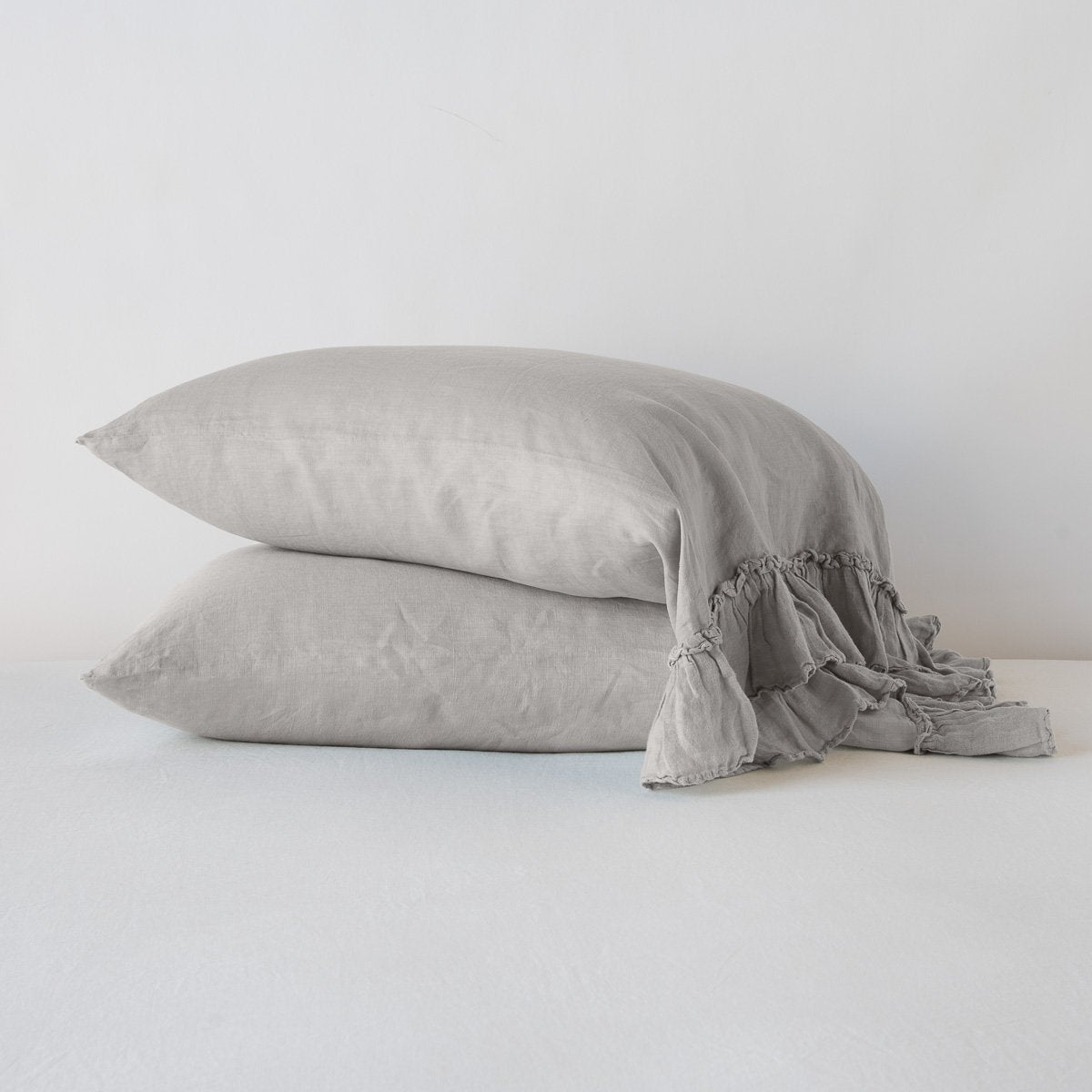 : Two Linen Whisper sleeping pillows, stacked at a slight angle against a plain background, showcasing ruffle trim detail - fog, side view.