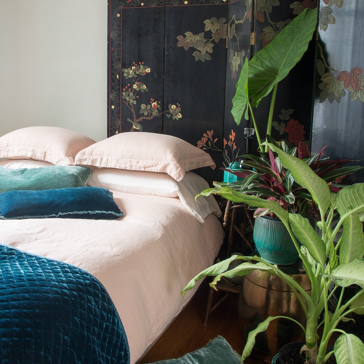 : Linen duvet cover in pearl on a neatly made bed. Silk velvet accents in blues and greens are complemented by on-tone ceramics, dark floral wall panel, and tall greenery - cropped three-quarter angle.