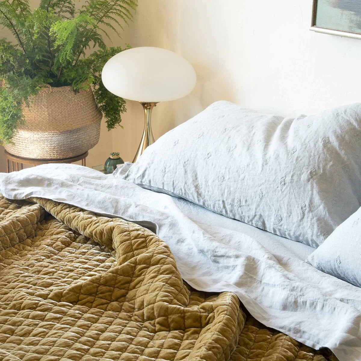 : Cloud linen sheeting with embroidered linen shams and rich gold toned silk velvet quilted coverlet, styled with a green fern in a woven basket and gold metal accents - cropped three-quarter angle.