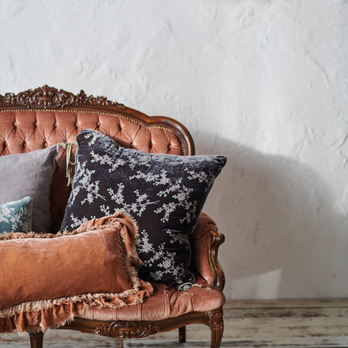 : Lynette 24 by 24 in moonlight, layered with grey, blue, and pink throw pillows on an antique tufted loveseat.