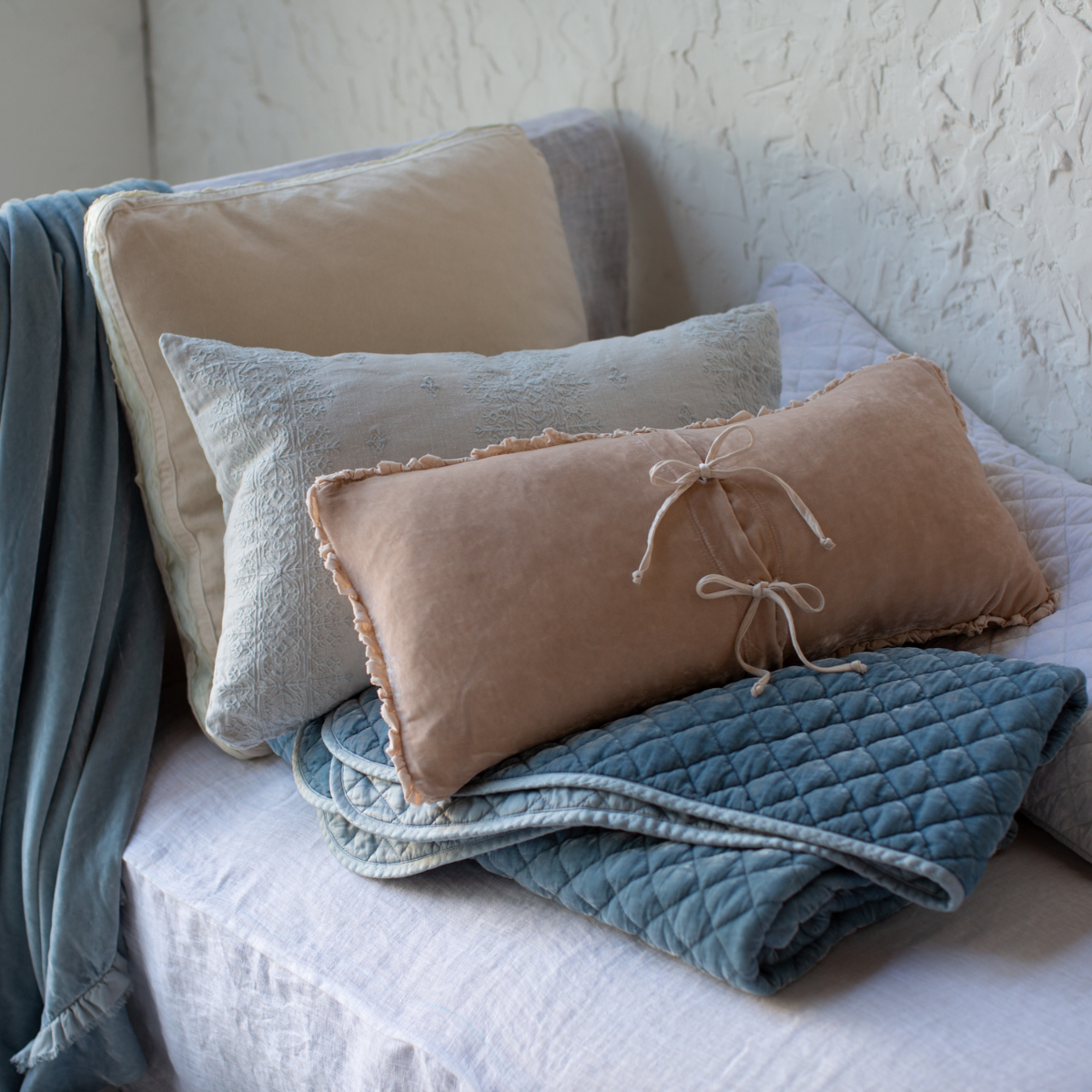 : Silk velvet throw blanket in cloud, neatly folded and layered with pink, cream and blue accessory pieces on a white linen daybed - cropped three quarter angle.