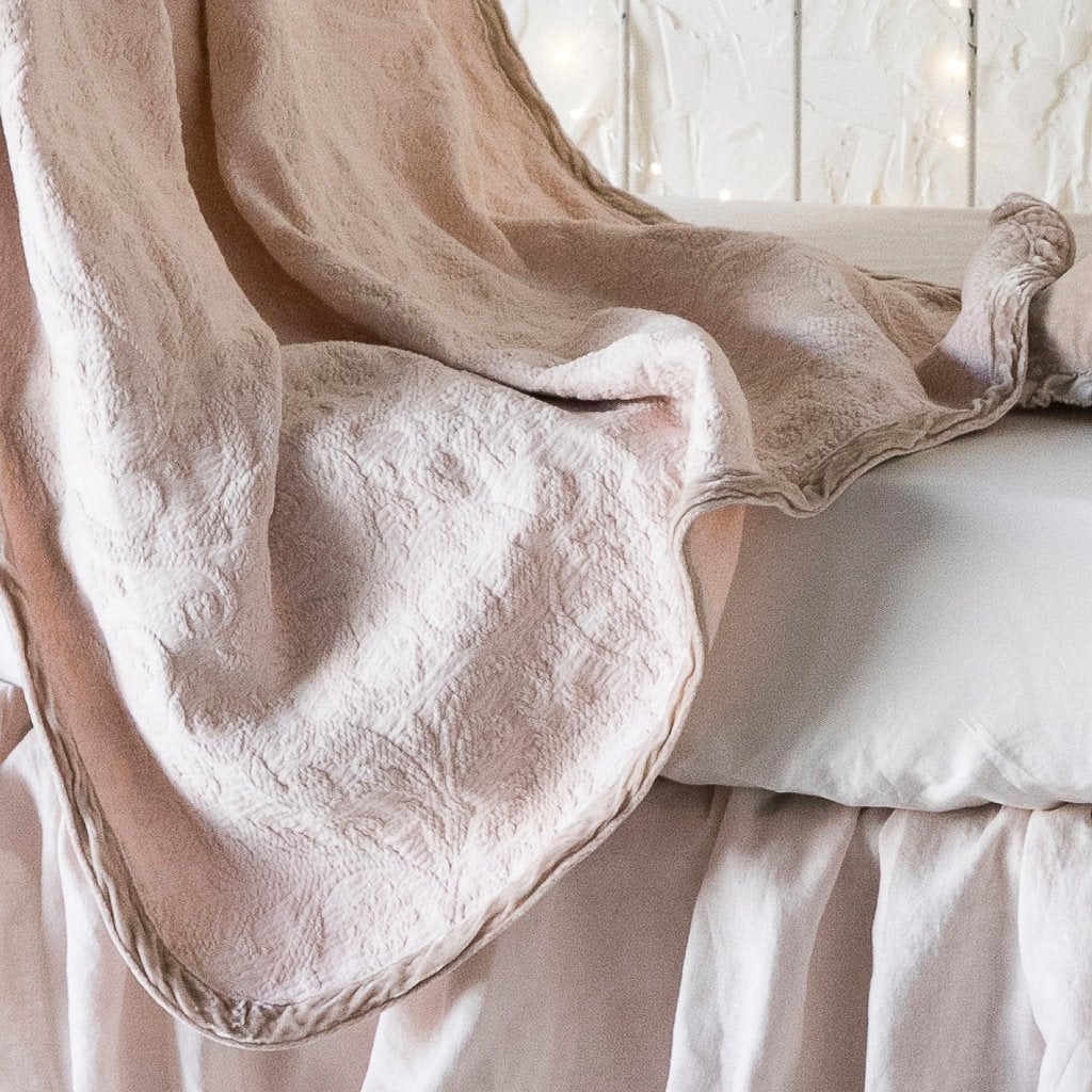 Pearl: A close up of the Adele baby blanket in pearl draped over a crib mattress, highlighting the texture of the cotton damask and the silk velvet edging, which absorbs the dye more richly. 