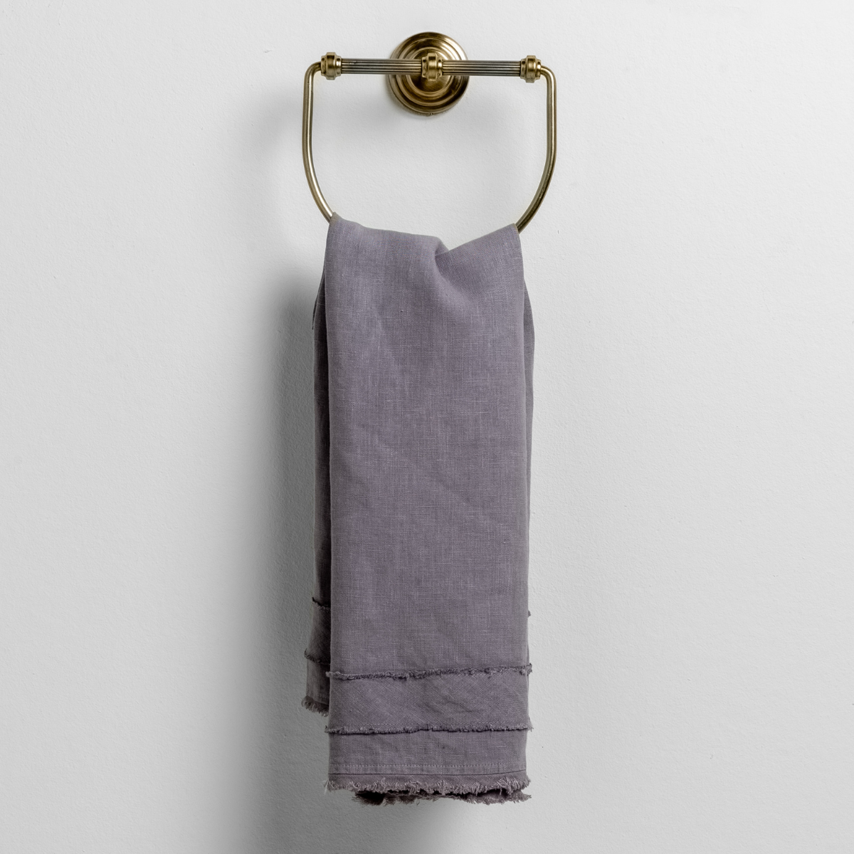 French Lavender: midweight linen guest towel with raw edge band at both ends hanging from a towel ring mounted to a white wall. 