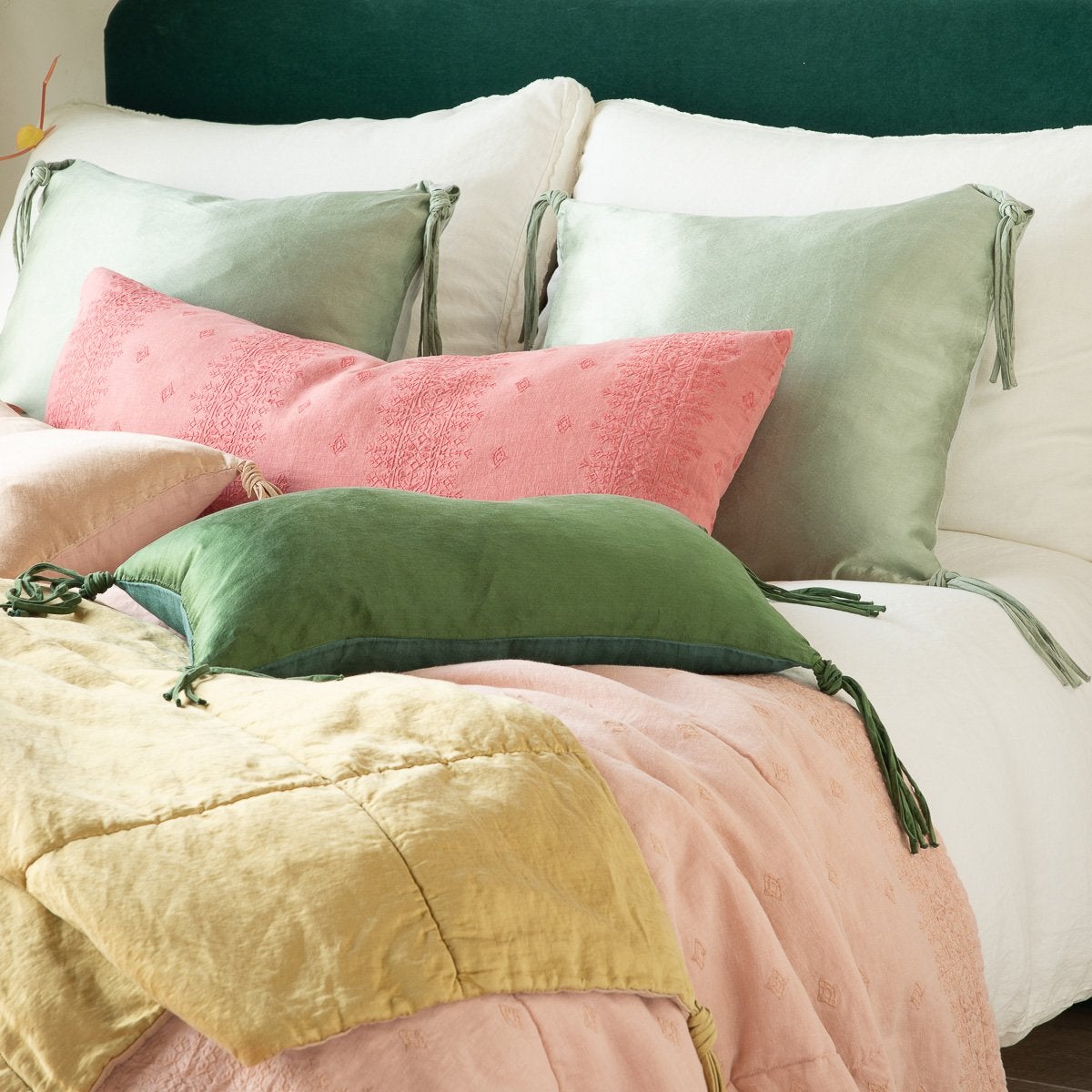 : Taline pillows and blanket in green and gold tones shine against pink-toned embroidered linen - cropped three-quarter angle.