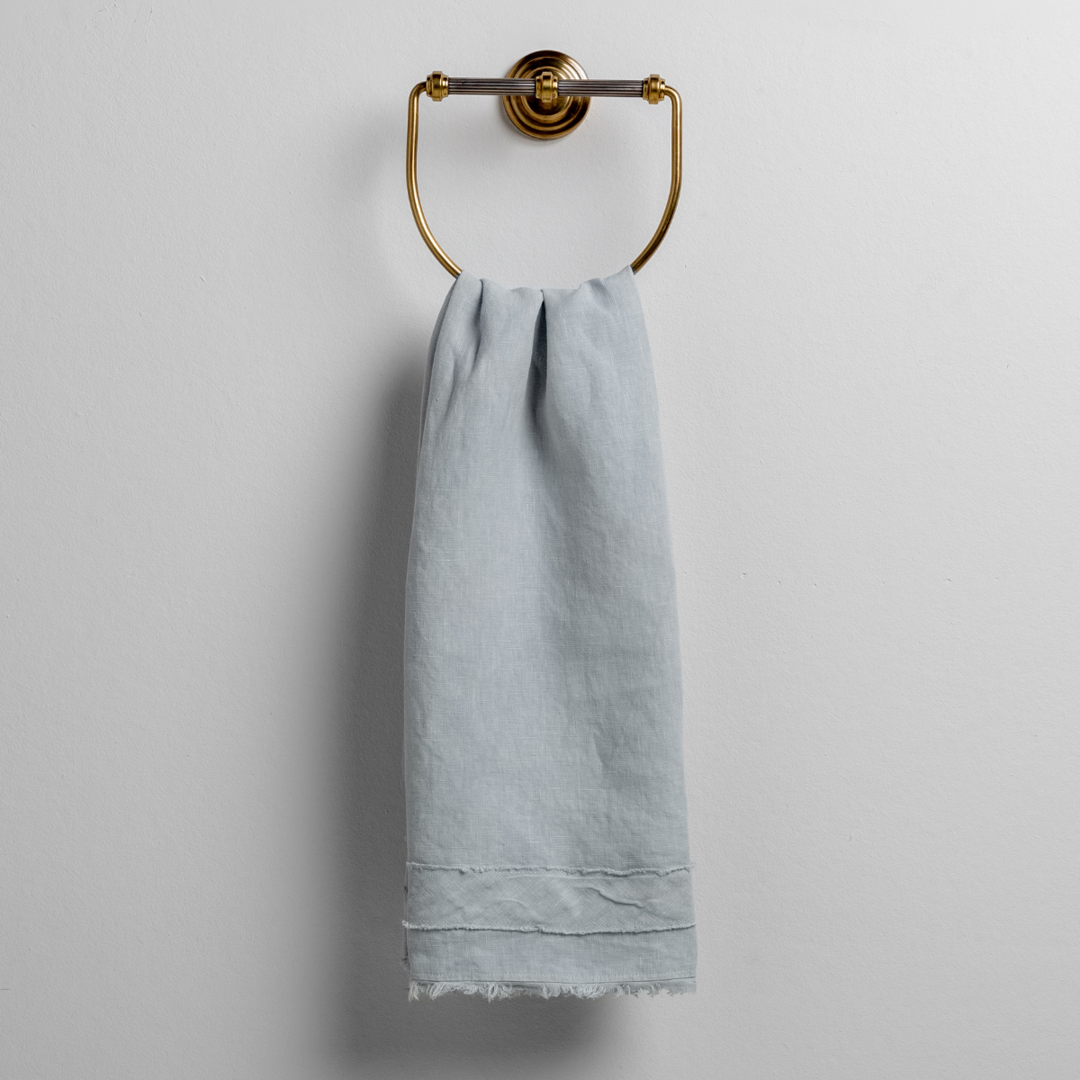 Cloud: midweight linen guest towel with raw edge band at both ends hanging from a towel ring mounted to a white wall. 