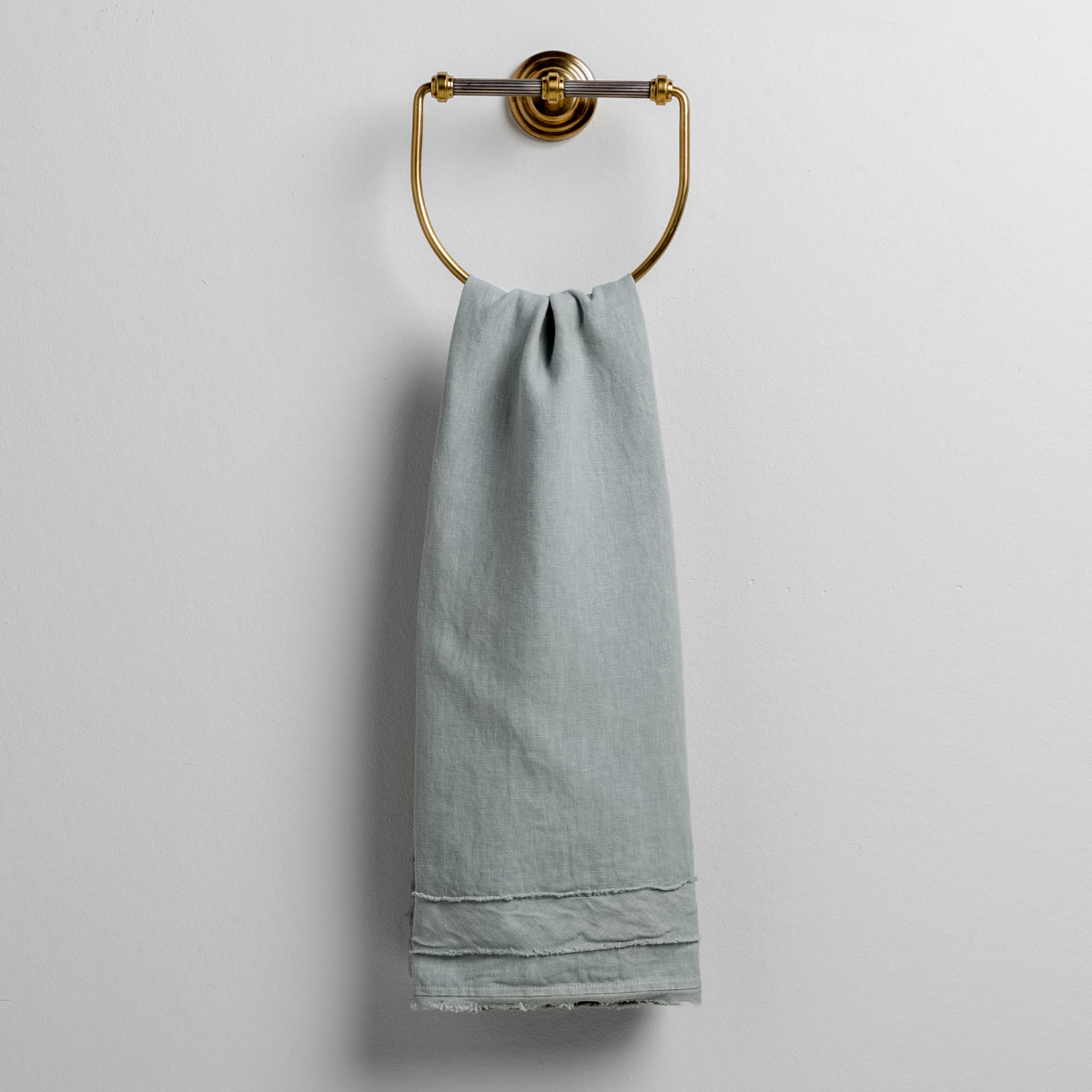 Eucalyptus: midweight linen guest towel with raw edge band at both ends hanging from a towel ring mounted to a white wall. 