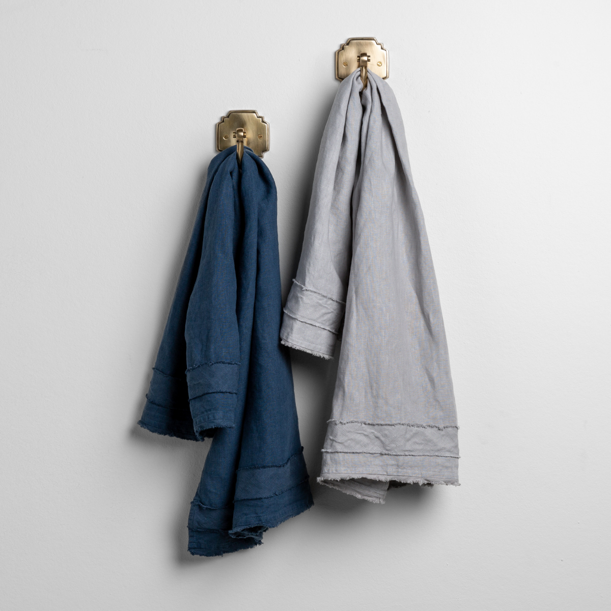 [allvariants]: pair of midweight linen guest towels in midnight and minderal on gold hooks against a white wall. 