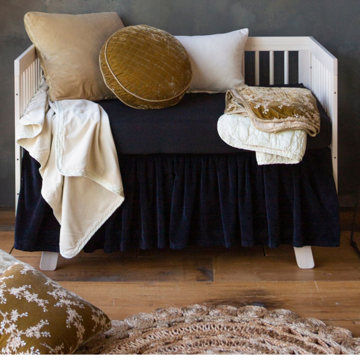 : black crib skirt and crib sheet on a neutral crib with neutral and golden toned blankets and throw pillows. 