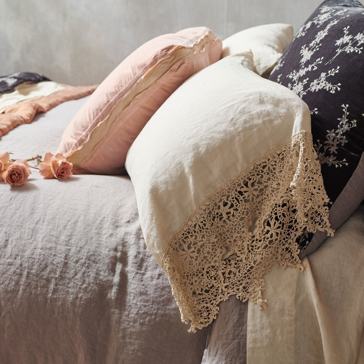 [allvariants]: Frida pillowcase in parchment layered on a bed with moonlight and pearl tones in linen, cotton velvet, and silk velvet - side view.