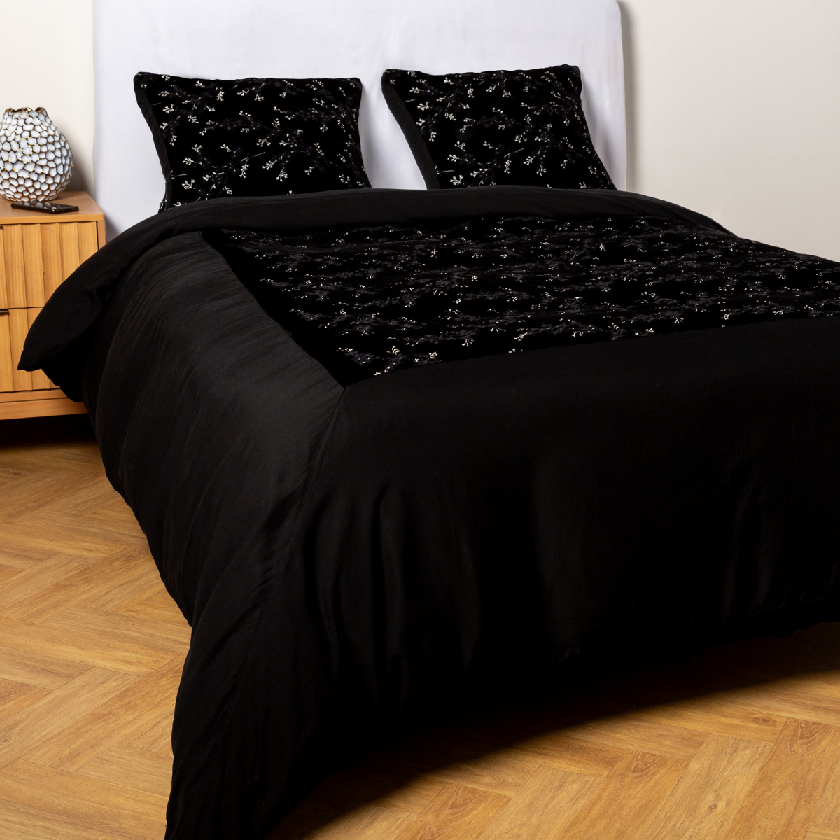 Corvino: A ¾ angle shot of an embroidered silk velvet and tencel duvet cover and matching shams with a vase and cell phone on the side table. 