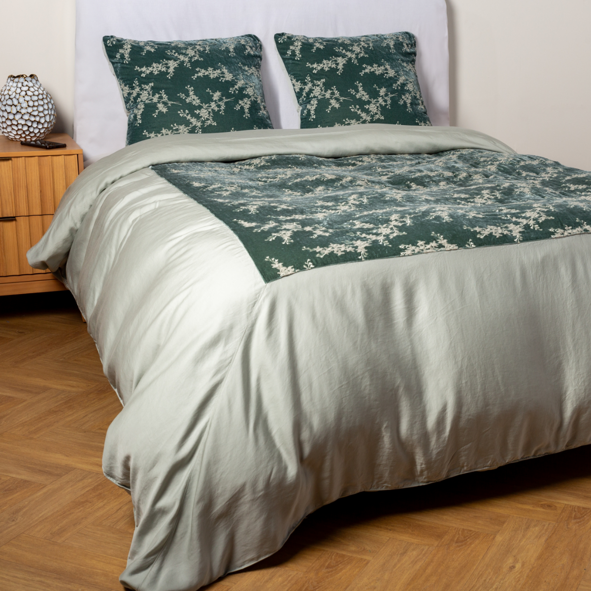 Eucalyptus: A ¾ angle shot of an embroidered silk velvet and tencel duvet cover and matching shams with a vase and cell phone on the side table. 