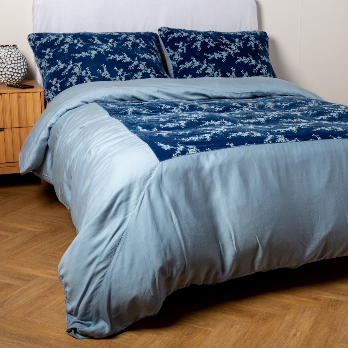 Tahoe Blue: A ¾ angle shot of an embroidered silk velvet and tencel duvet cover and matching shams with a vase and cell phone on the side table. 