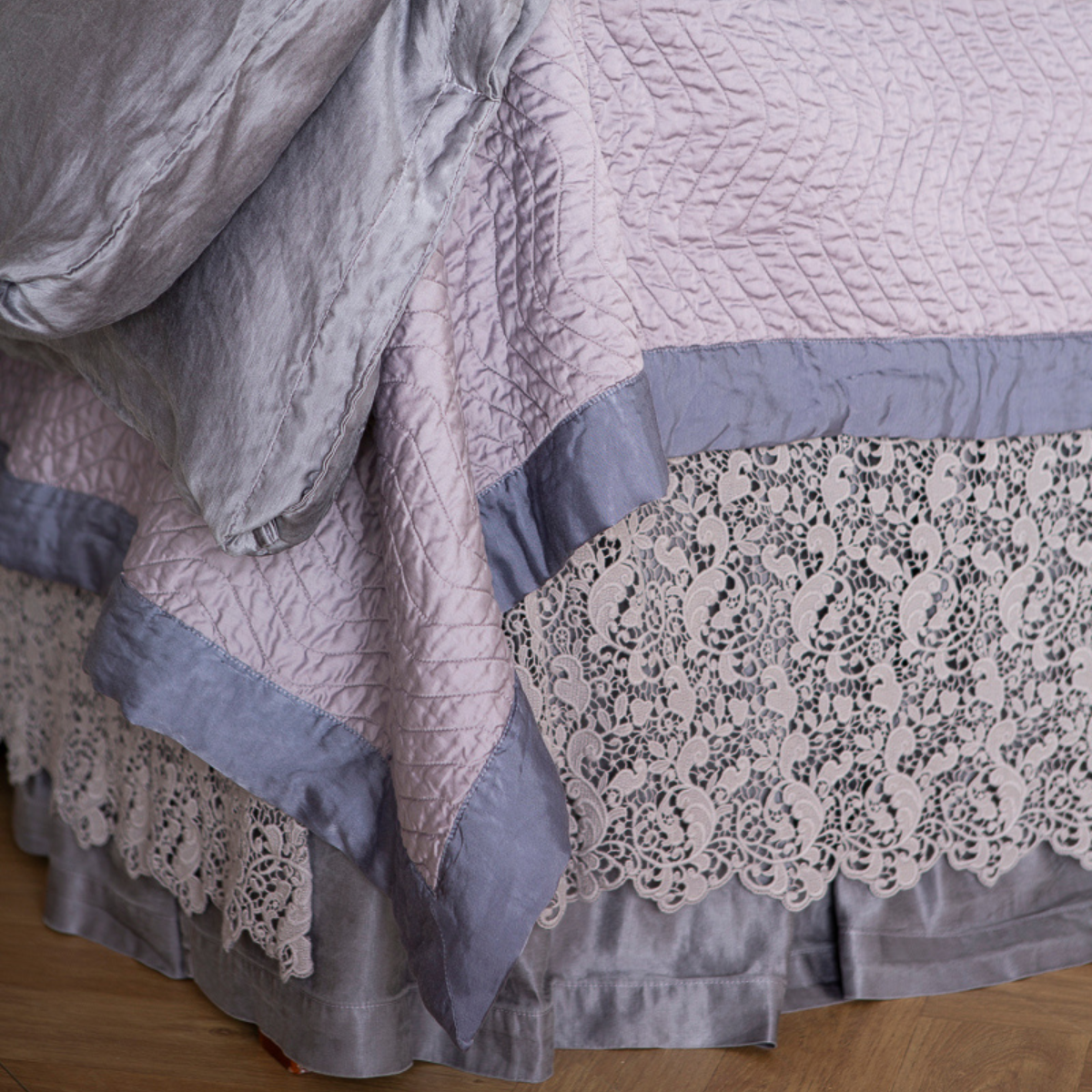 : the base and corner of a monochromatic bed in lavender tones showing a duvet cover and coverlet layered with a cotton lace bed skirt over a charmeuse skirt. 