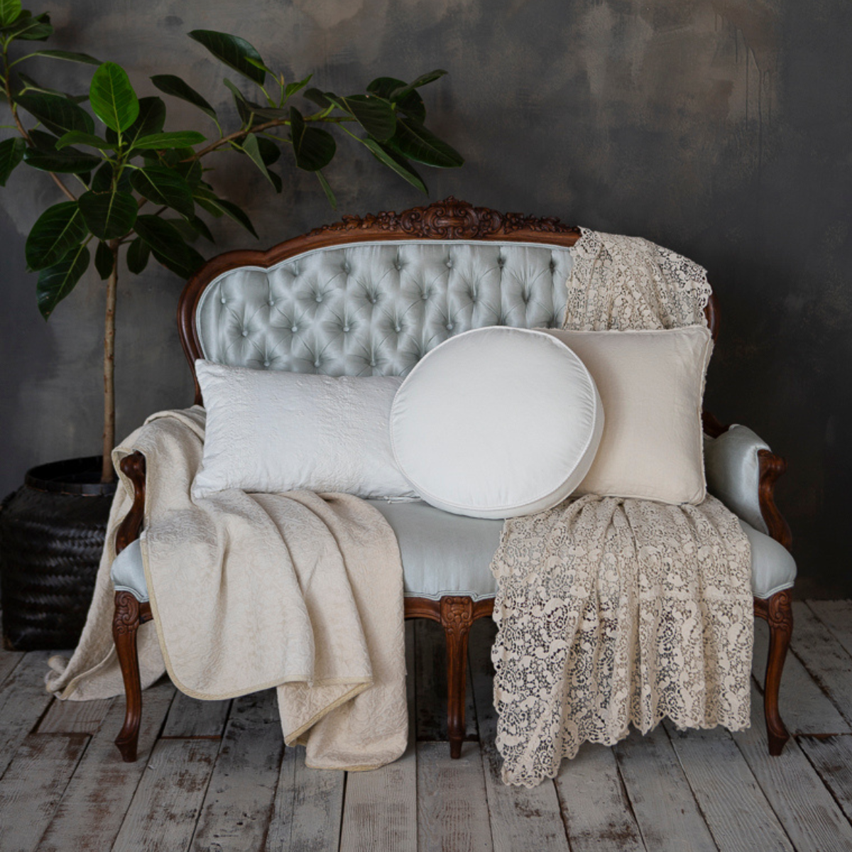 : a lace bed scarf draped on a sofa with throw pillows and a throw blanket in neutral tones on a light blue sofa with wooden details on its frame. 