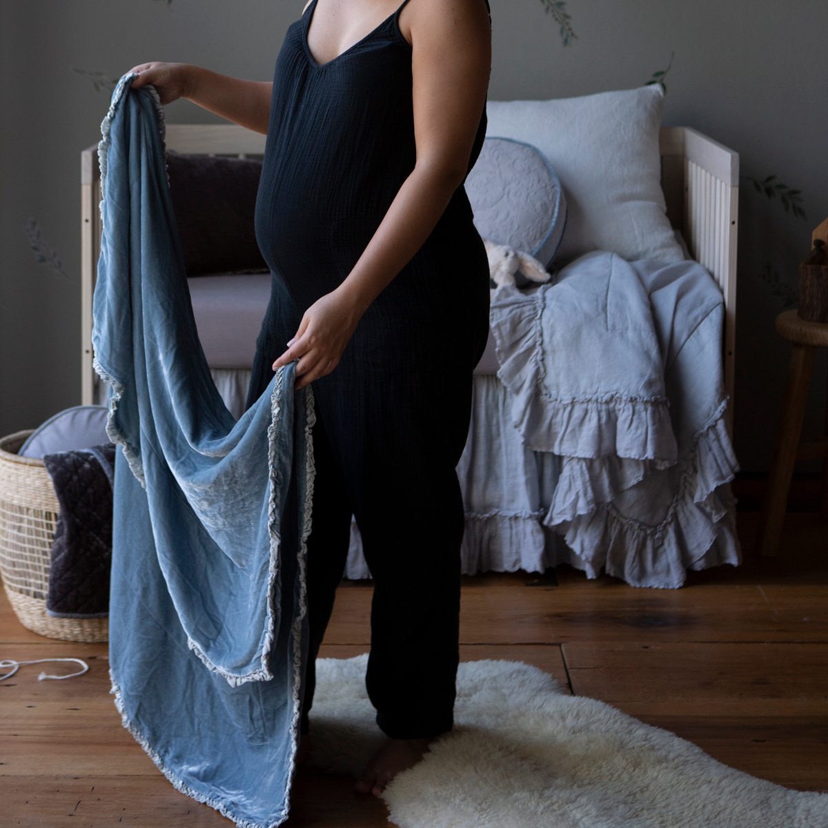 [allvariants]: A pregnant woman, seen from the shoulders down, holding a blanket open in cloud in a blue and grey toned nursery. 