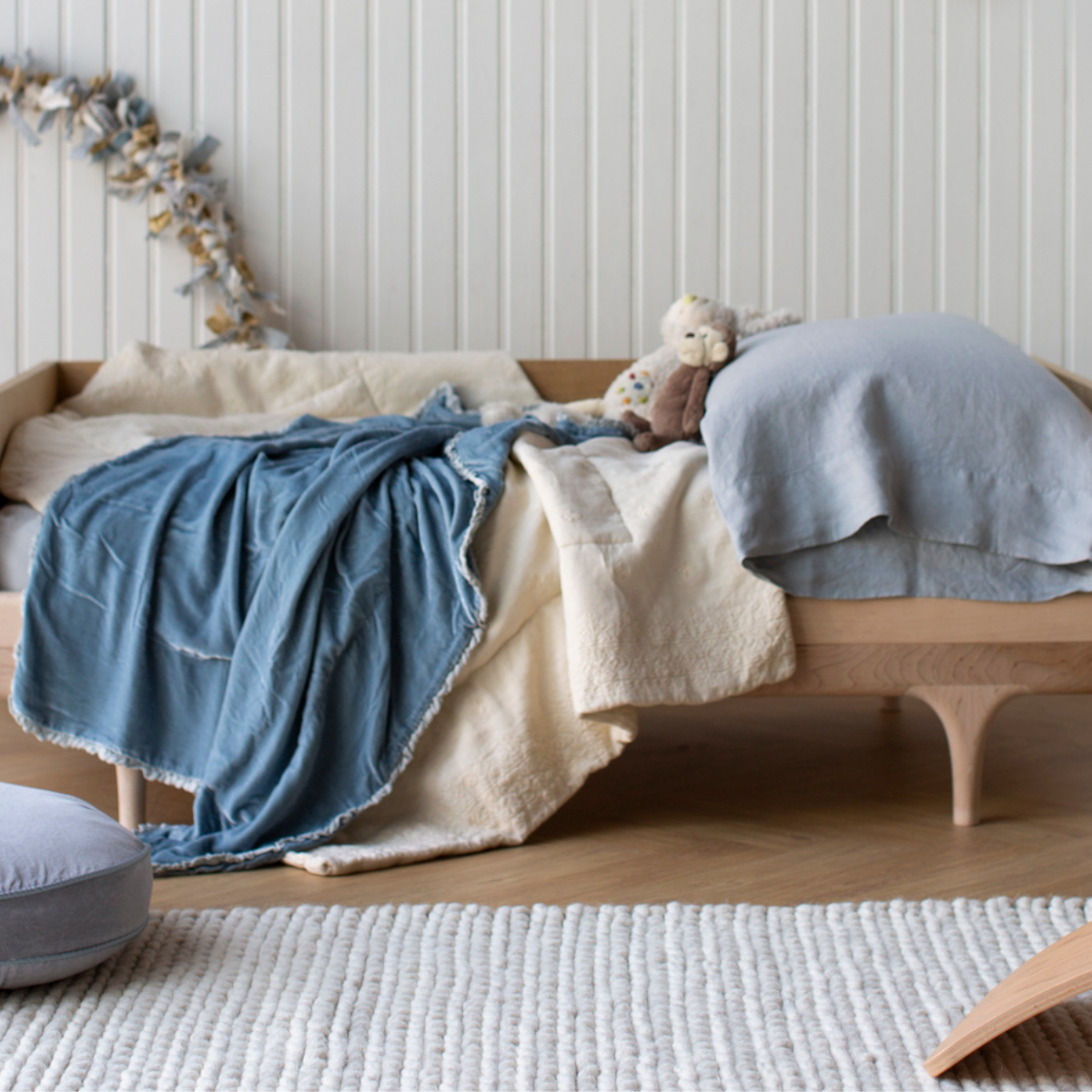 : Rumpled blanket in cloud layered over a toddler bed in cream and blue grey tones, in a bright nursery with warm wood accents.