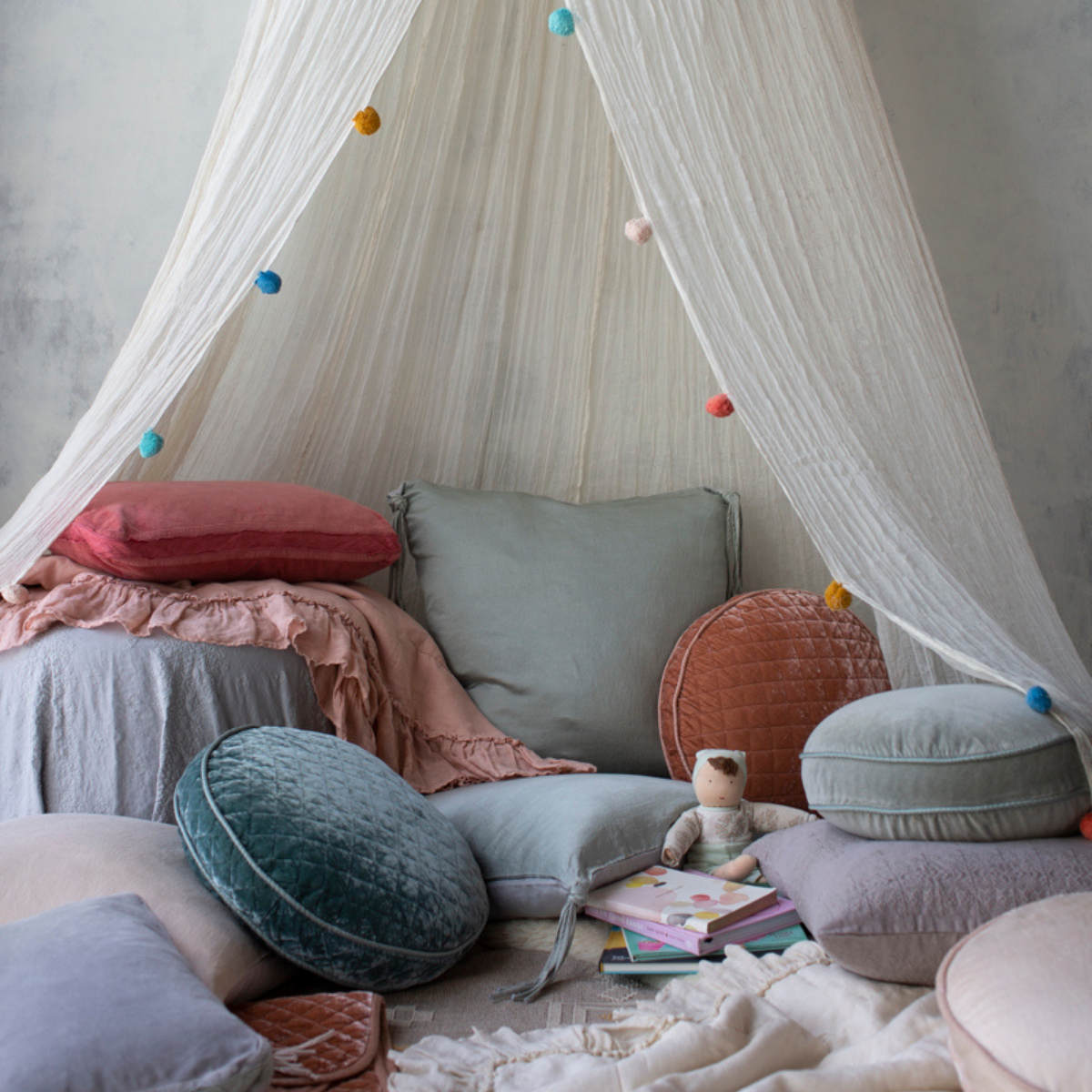 : Blankets in rouge and parchment rumpled among a canopy-covered nook on the floor covered witha cozy mix of pillows, blankets, books and a doll. 