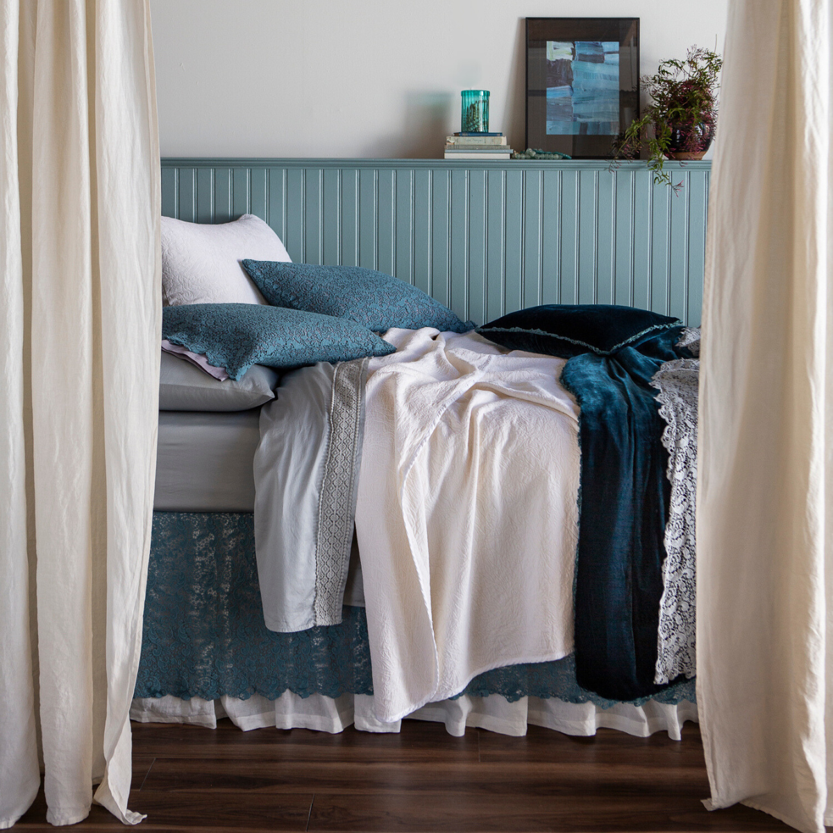 [allvariants]: side view of a bed dressed with lace pillowcases, flat sheet, bed skirt and bed scarf layered with cotton sateen sheets, chenille and cotton jacquard bedspread and shams and silk velvet accent pillow and bed end blanket — shown from the side with curtain panels framing the bed that's against a light blue paneled wall. 