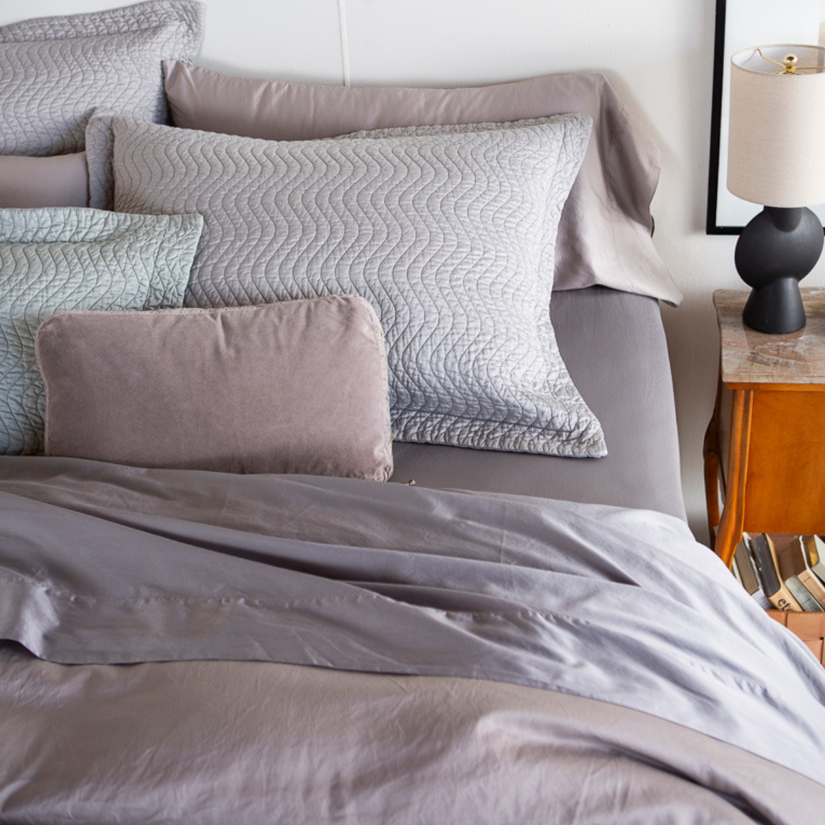 : a slightly overhead angle of the head of a bed with quilted cotton sateen shams, throw pillows and unquilted cotton sateen sleeping pillows. The colors are cool and tonal all in different types of cotton. 