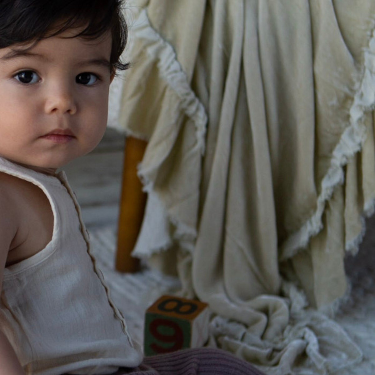 : silk velvet blanket in parchment cascading down the front of a chair in the background shows the drape of the fabric and trim detail. A young child is looking at the camera while playing with blocks. 