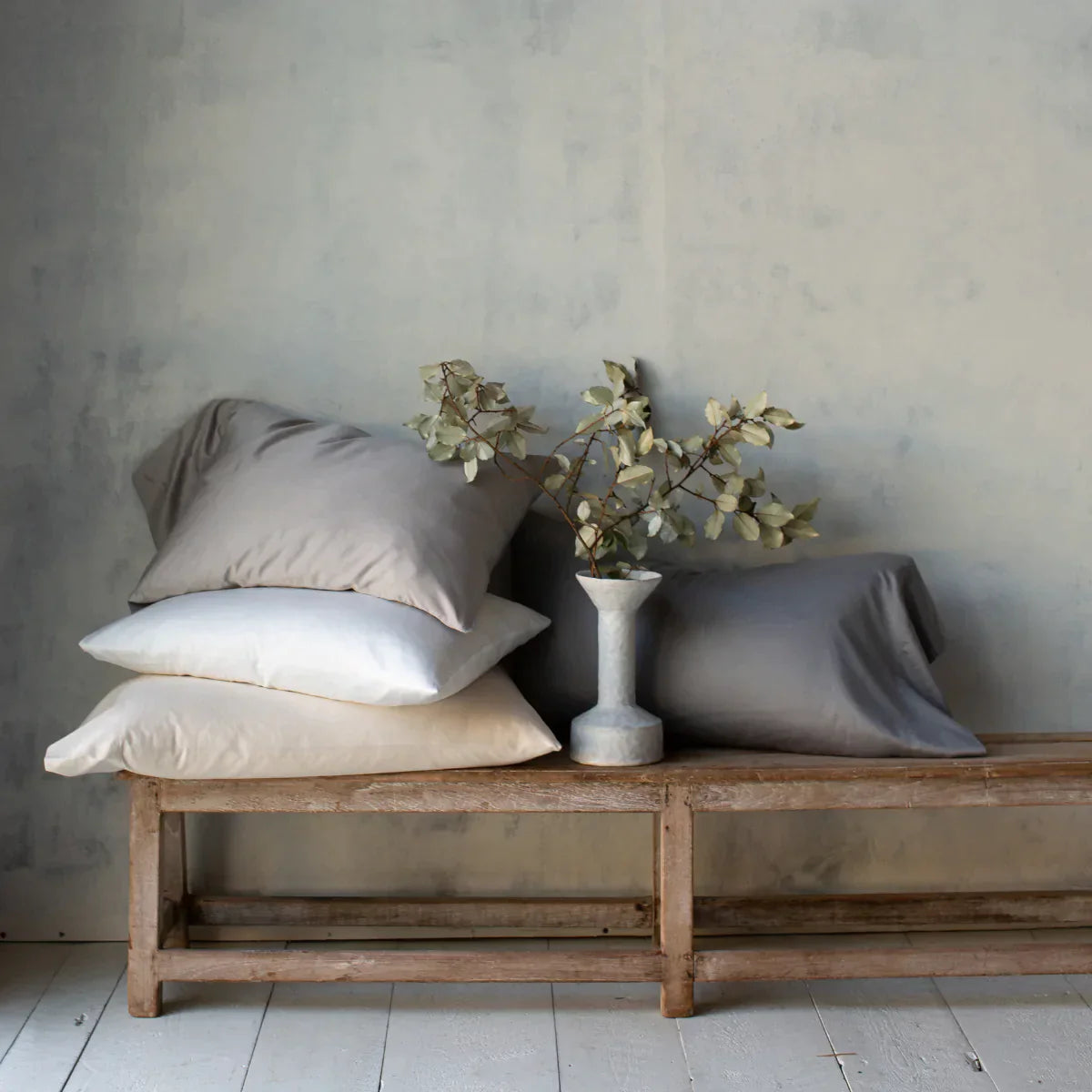 : Bria sleeping pillows in various white and grey tones, stacked on a wooden bench against a textured neutral wall. A ceramic vase with greenery sits between the pillows.