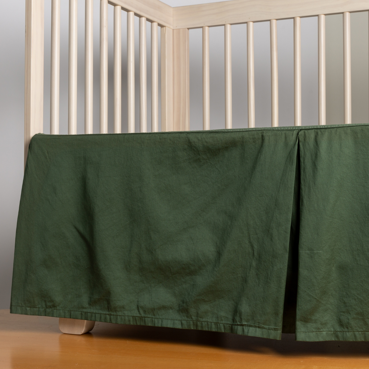 Juniper: cotton sateen crib skir tiwth a center pleat shown straight on from a slight angle in a crib without a crib mattress. 
