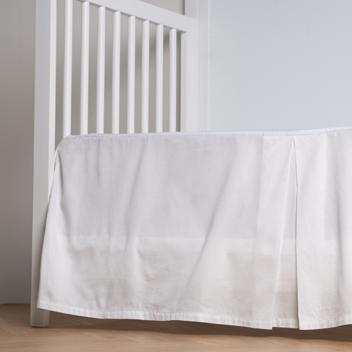 Winter White: cotton sateen cribi skirt with a center pleat shown straight on from a slight angle in a crib without a crib mattress. 