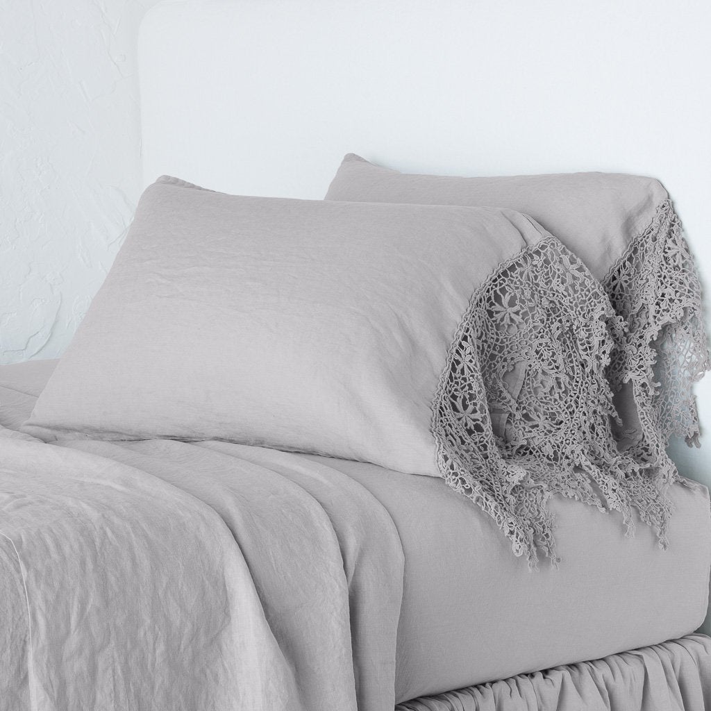 Fog: Lace trimmed linen pillowcases leaning against a plain wall on a monochromatic bed - cropped three-quarter angle.