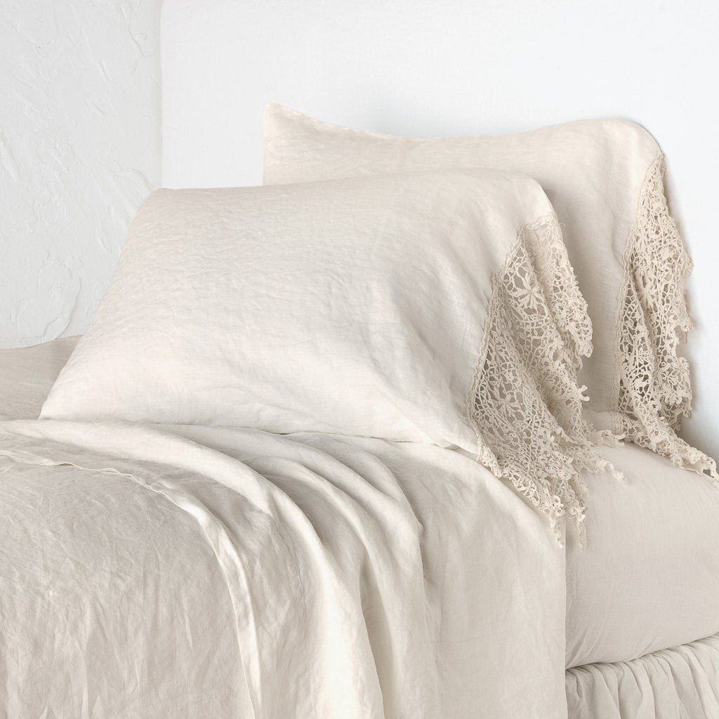 Parchment: Lace trimmed linen pillowcases leaning against a plain wall on a monochromatic bed - cropped three-quarter angle.