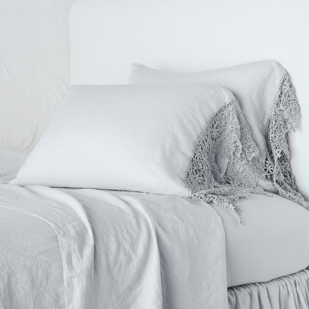 Sterling: Lace trimmed linen pillowcases leaning against a plain wall on a monochromatic bed - cropped three-quarter angle.