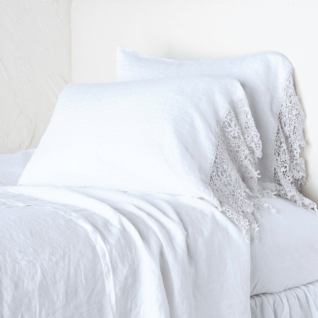 White: Lace trimmed linen pillowcases leaning against a plain wall on a monochromatic white bed - cropped three-quarter angle.