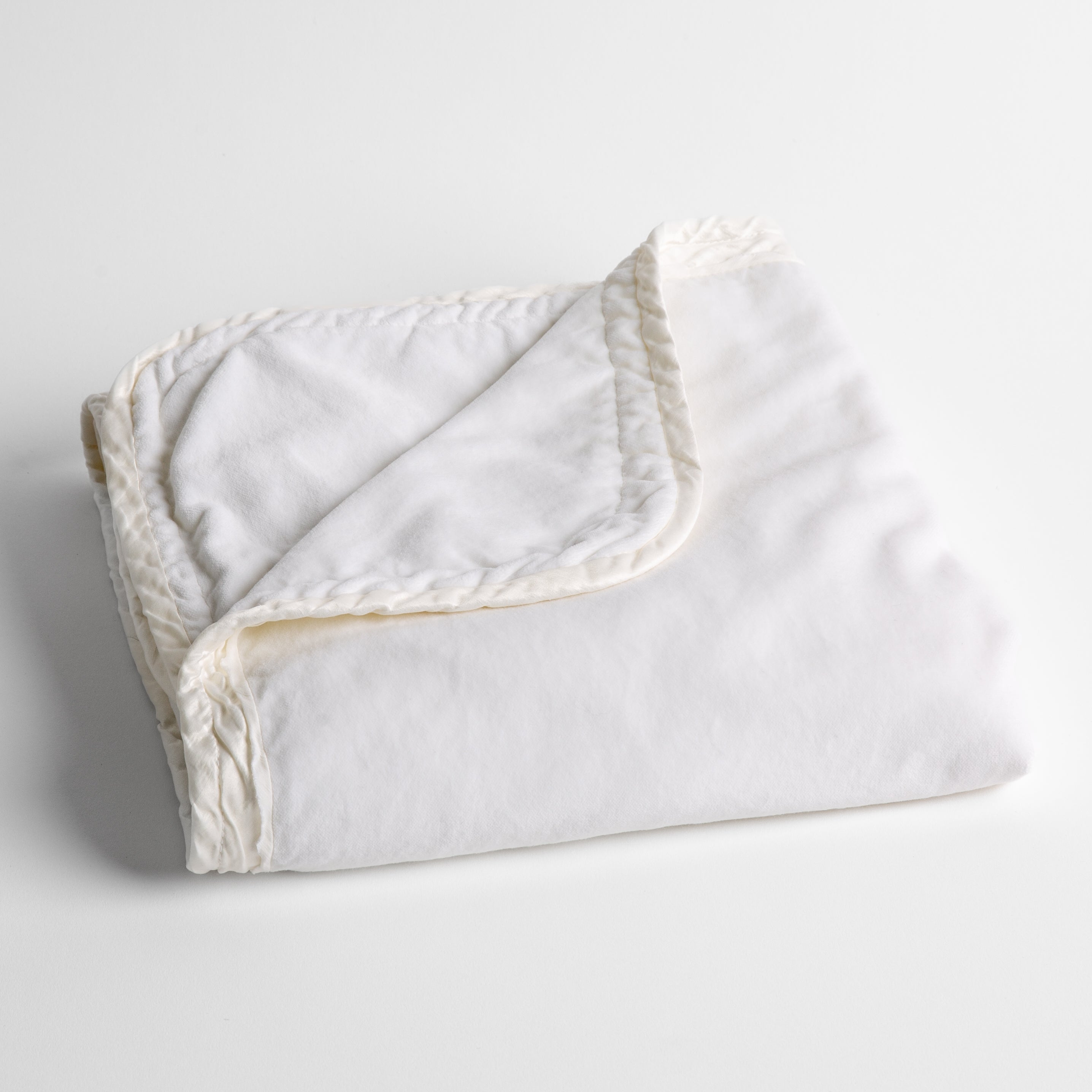 White: cotton velvet baby blanket trimmed with charmeuse, folded with a covern folded back to show trim contrast; shot overhead and at a slight angle against a white background. 