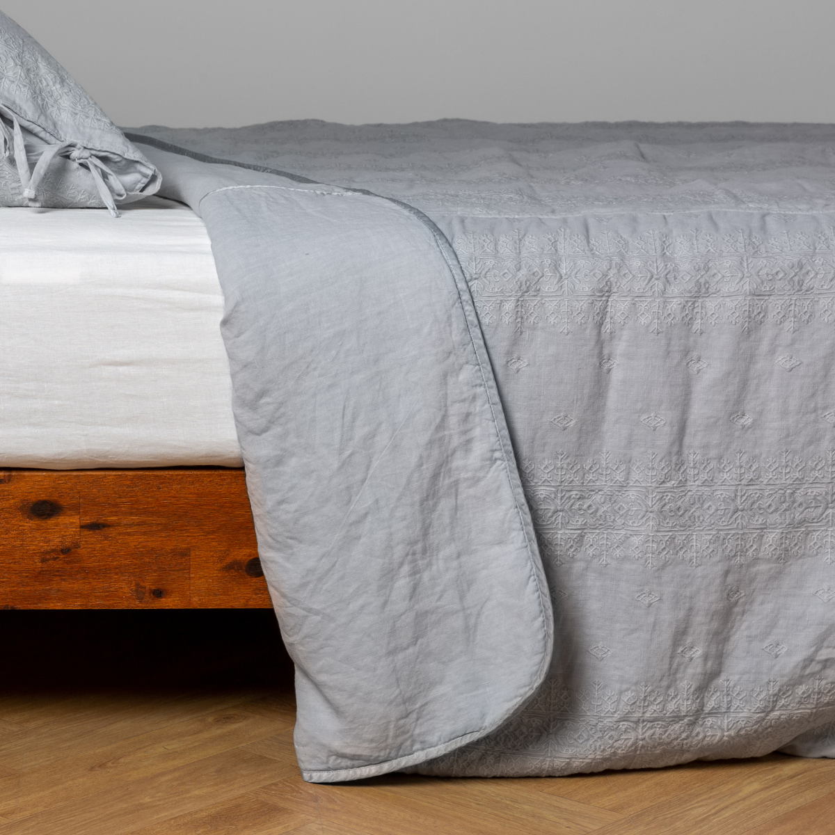 Cloud: White: embroidered bedspread with corner pulled back  showing the plain linen back. Shown with a white fitted sheet and matching sham, the bedframe is slightly visible  and the bed is against a white wall and on a medium wood floor. 