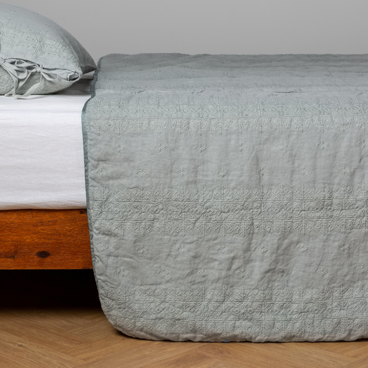 Eucalyptus:  embroidered midweight linen bedspread with matching sham on a bed with a white fitted sheet. Shot from the side view, the bedframe is slightly visible  against a white background and medium wood floor. 