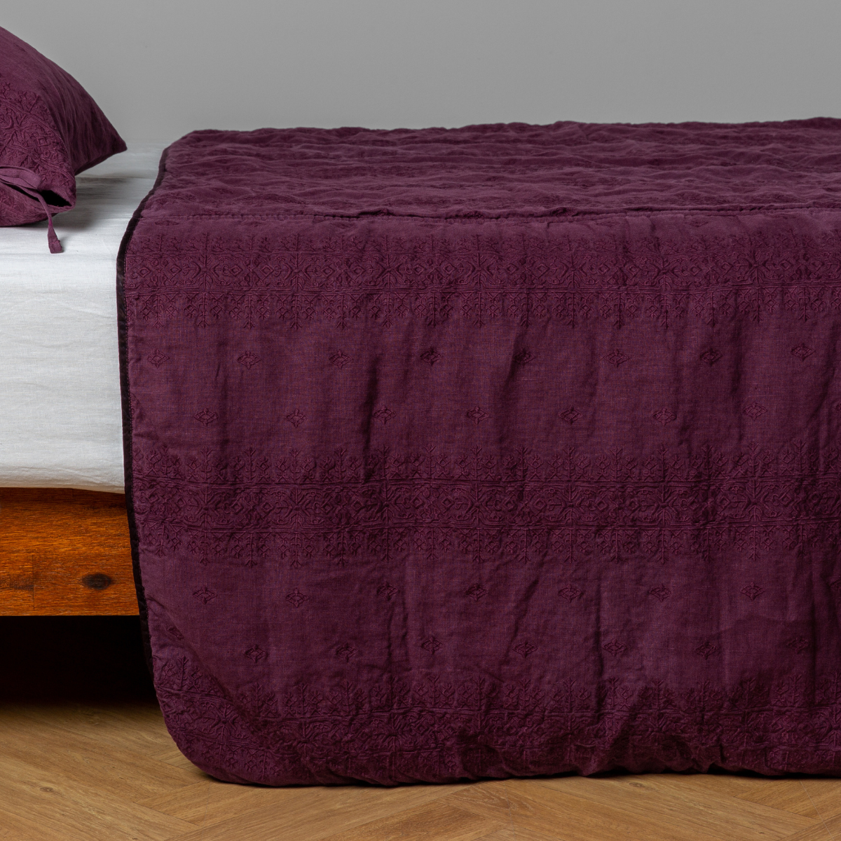 Fig: embroidered midweight linen bedspread with matching sham on a bed with a white fitted sheet. Shot from the side view, the bedframe is slightly visible  against a white background and medium wood floor. 