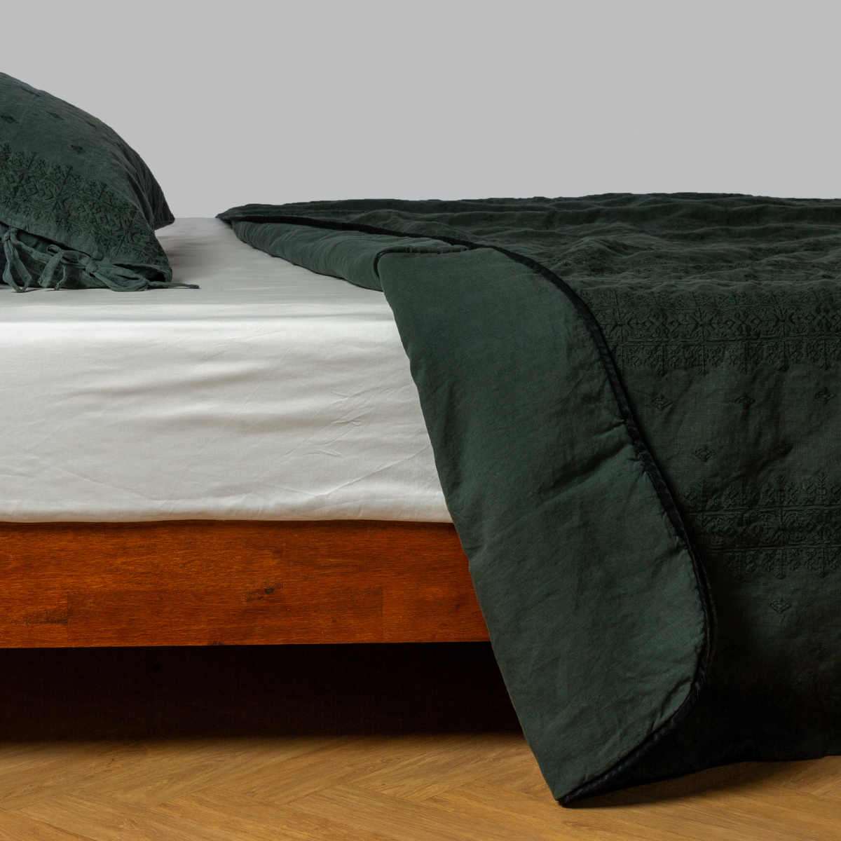 Juniper: embroidered midweight linen bedspread, corner folded back, with matching sham on a bed with a white fitted sheet. shot from the side view, the bedframe is slightly visible against a white background and medium wood floor. 