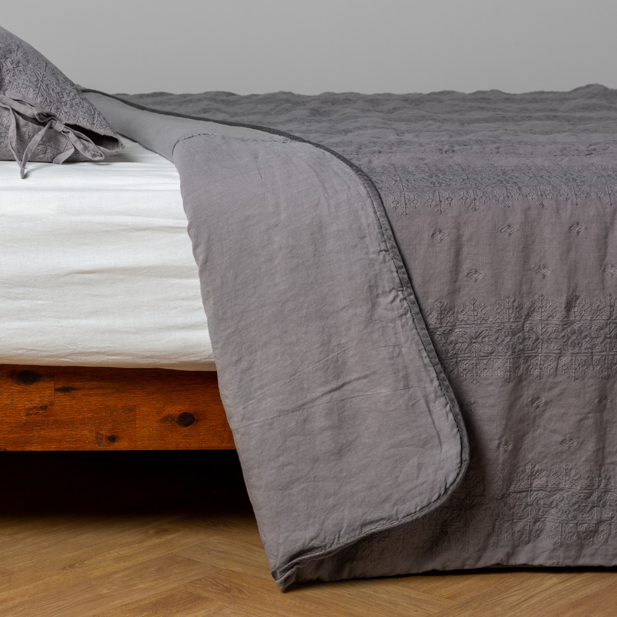 Moonlight: White: embroidered bedspread with corner pulled back  showing the plain linen back. Shown with a white fitted sheet and matching sham, the bedframe is slightly visible  and the bed is against a white wall and on a medium wood floor. 