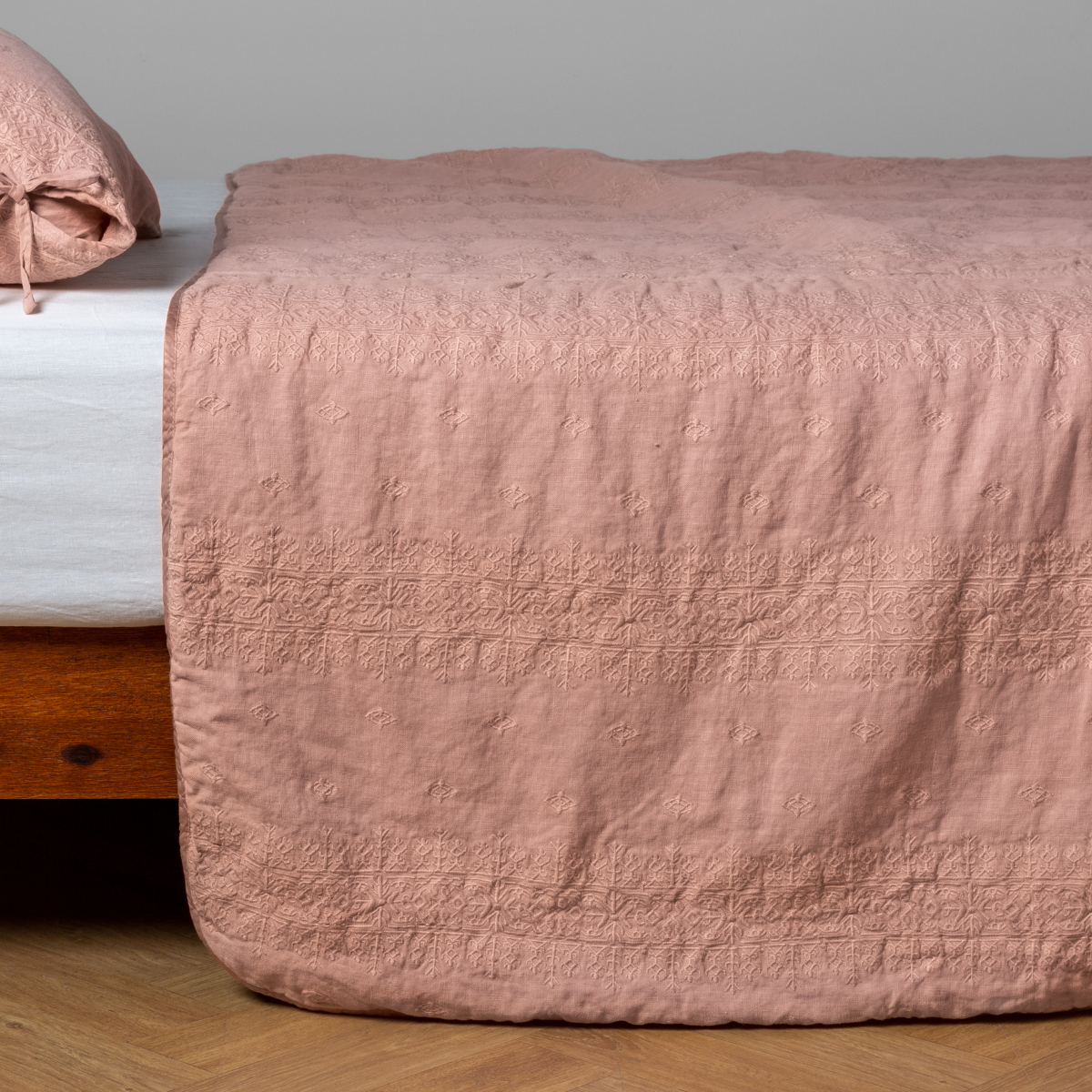 Rouge:  embroidered midweight linen bedspread with matching sham on a bed with a white fitted sheet. Shot from the side view, the bedframe is slightly visible  against a white background and medium wood floor. 