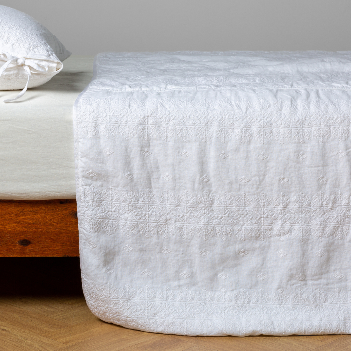 White: embroidered midweight linen bedspread with matching sham on a bed with a white fitted sheet. Shot from the side view, the bedframe is slightly visible  against a white background and medium wood floor. 