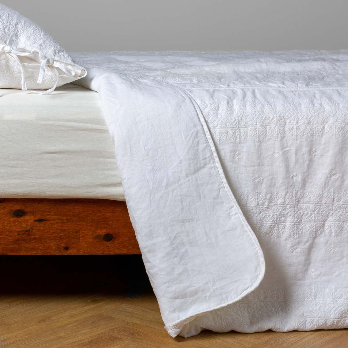 White: embroidered bedspread with corner pulled back  showing the plain linen back. Shown with a white fitted sheet and matching sham, the bedframe is slightly visible  and the bed is against a white wall and on a medium wood floor. 