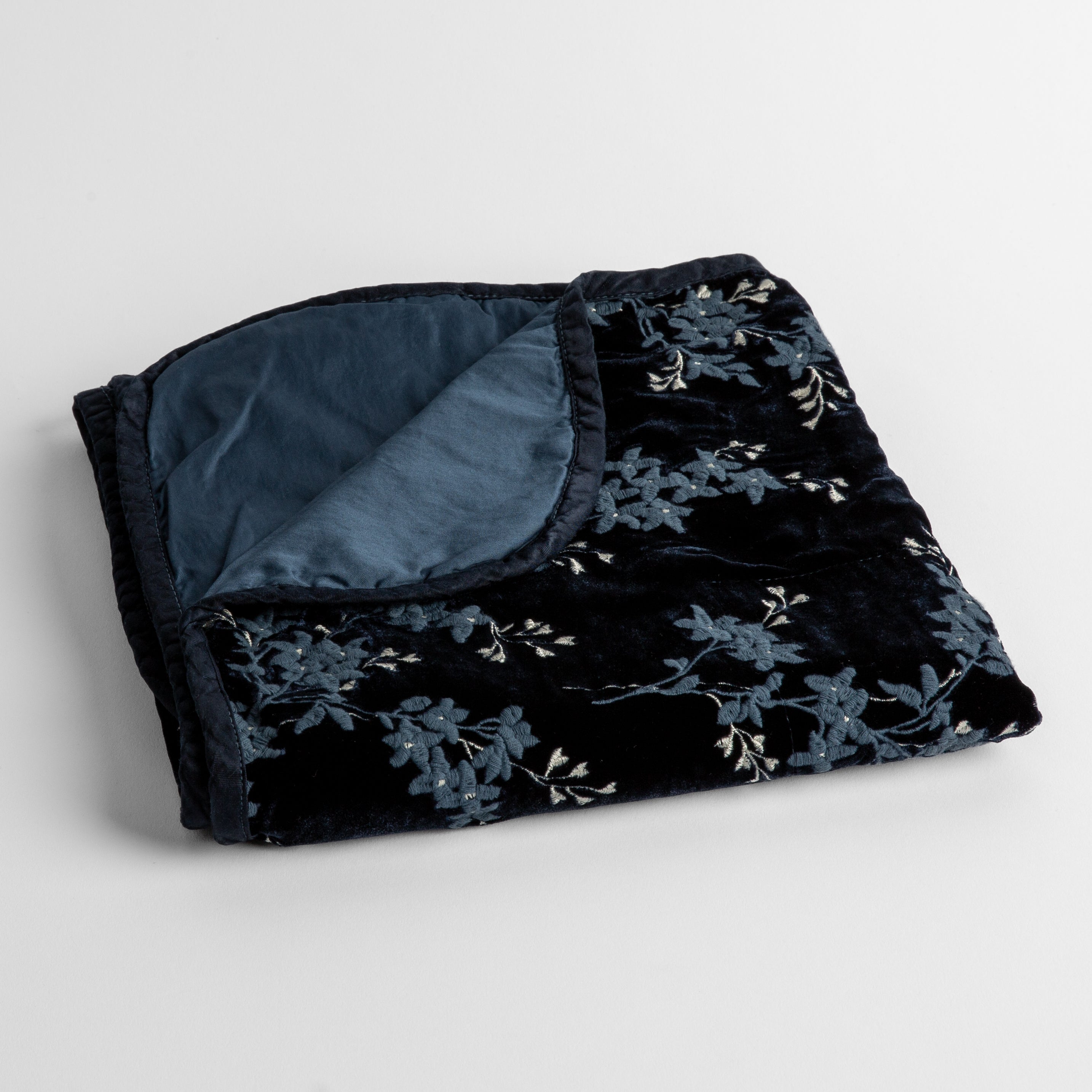 Midnight: embroidered silk velvet baby blanket folded with a corner folded back to show the trim and back of the blanket - shot overhead at a slight angle against a white background. 