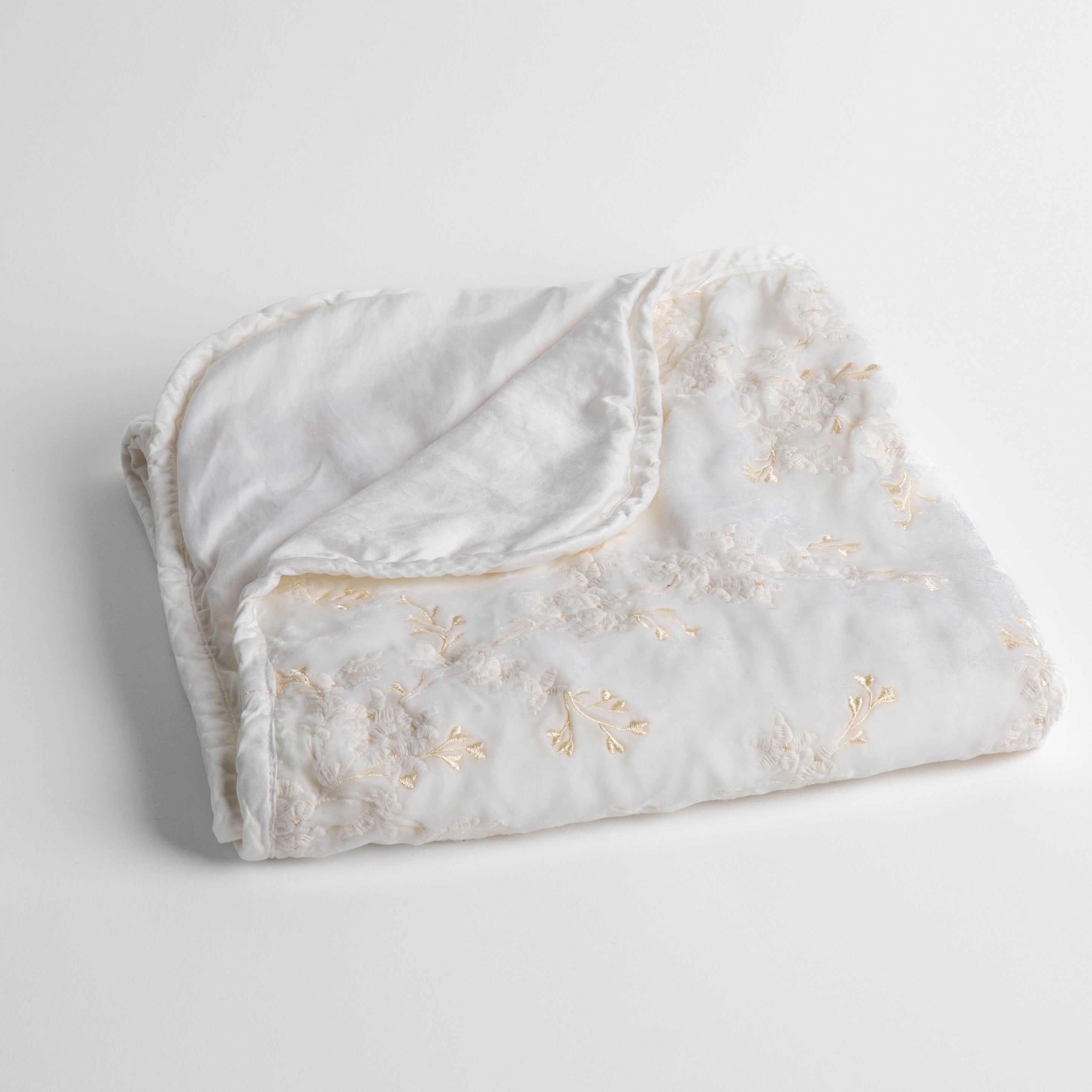 White: embroidered silk velvet baby blanket folded with a corner folded back to show the trim and back of the blanket - shot overhead at a slight angle against a white background. 