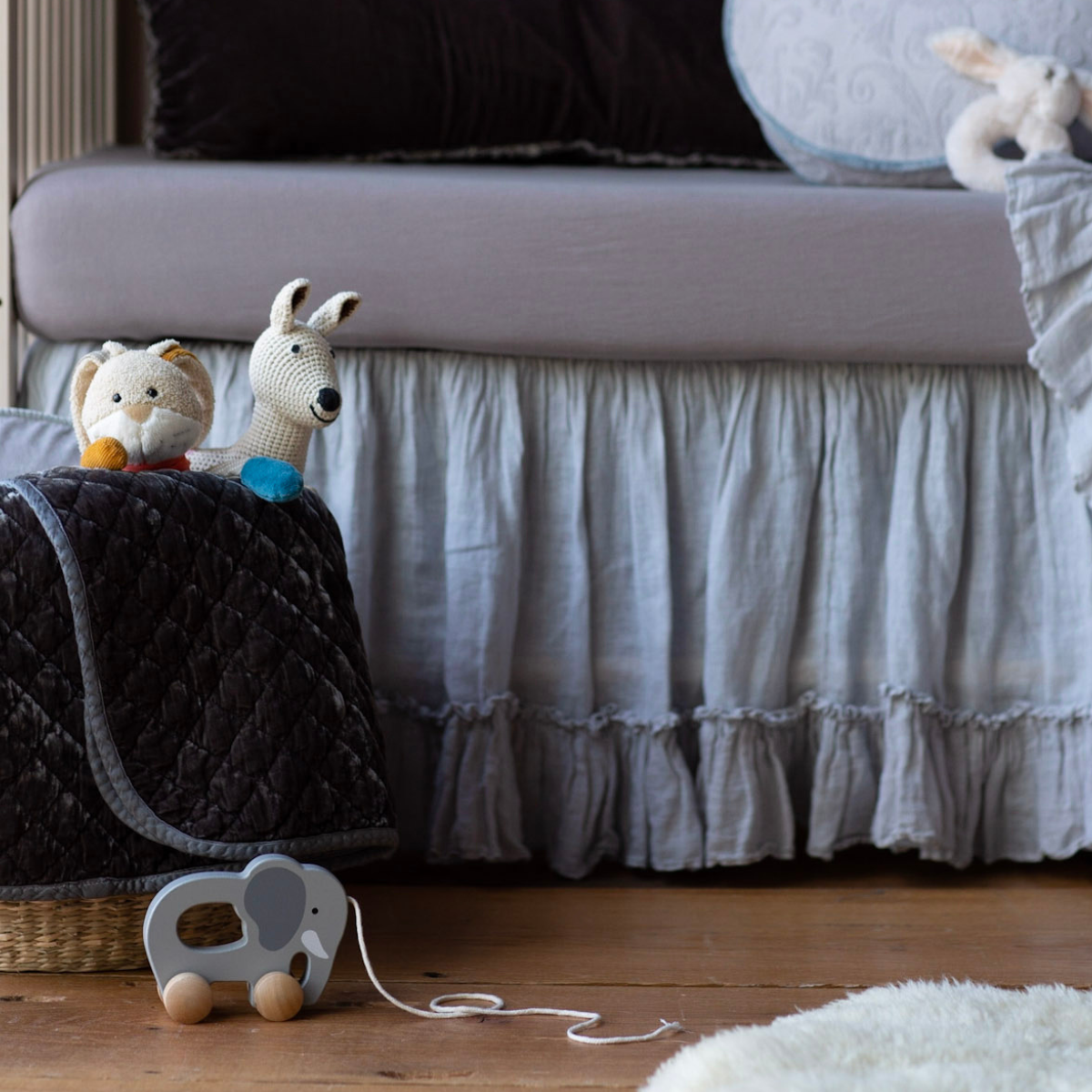 [allvariants]: Blanket in moonlight folded and draped over a basket in blue and grey nursery scene, shown with stuffed animals and wooden elephant pull toy.